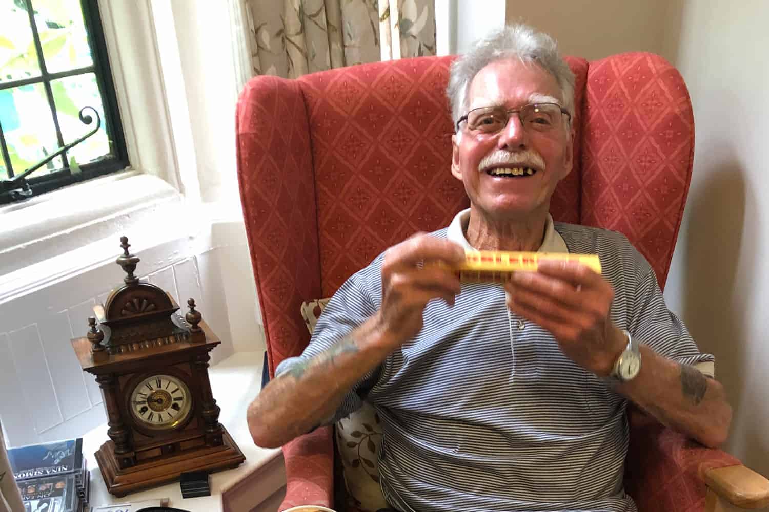 An elderly person with a bright smile sits in a patterned armchair, holding a small object related to their personalized healthcare plan, with a vintage mantle clock on a table beside them.