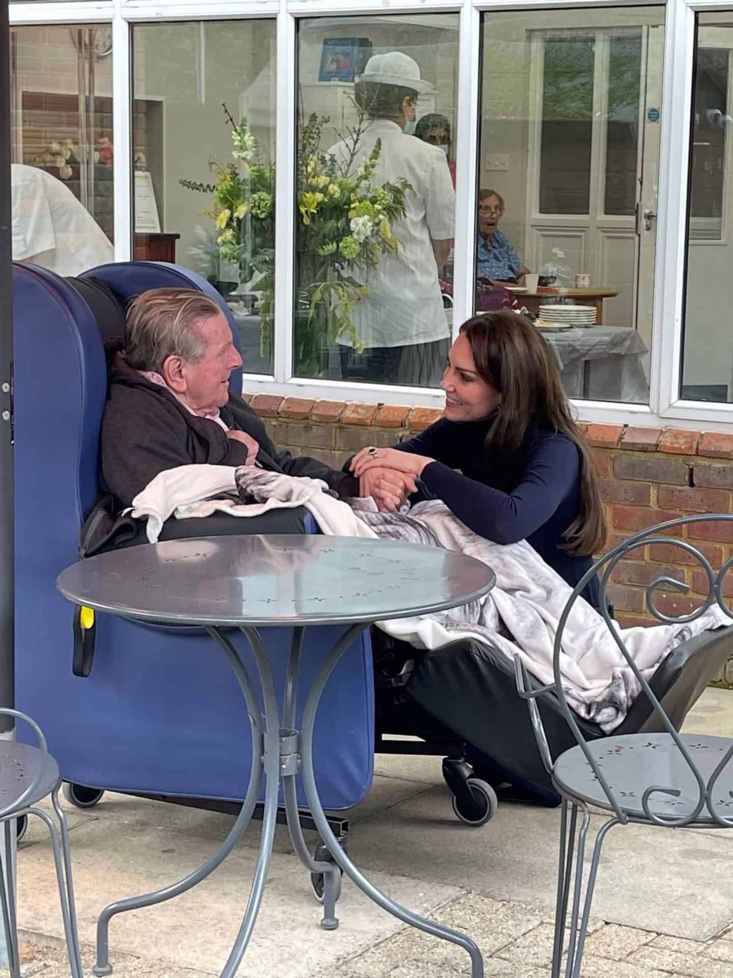 A tender moment between two individuals: an elderly person in a wheelchair and a woman in a navy top, holding hands and engaging in a caring conversation outdoors about choosing a nursing home, with a building and