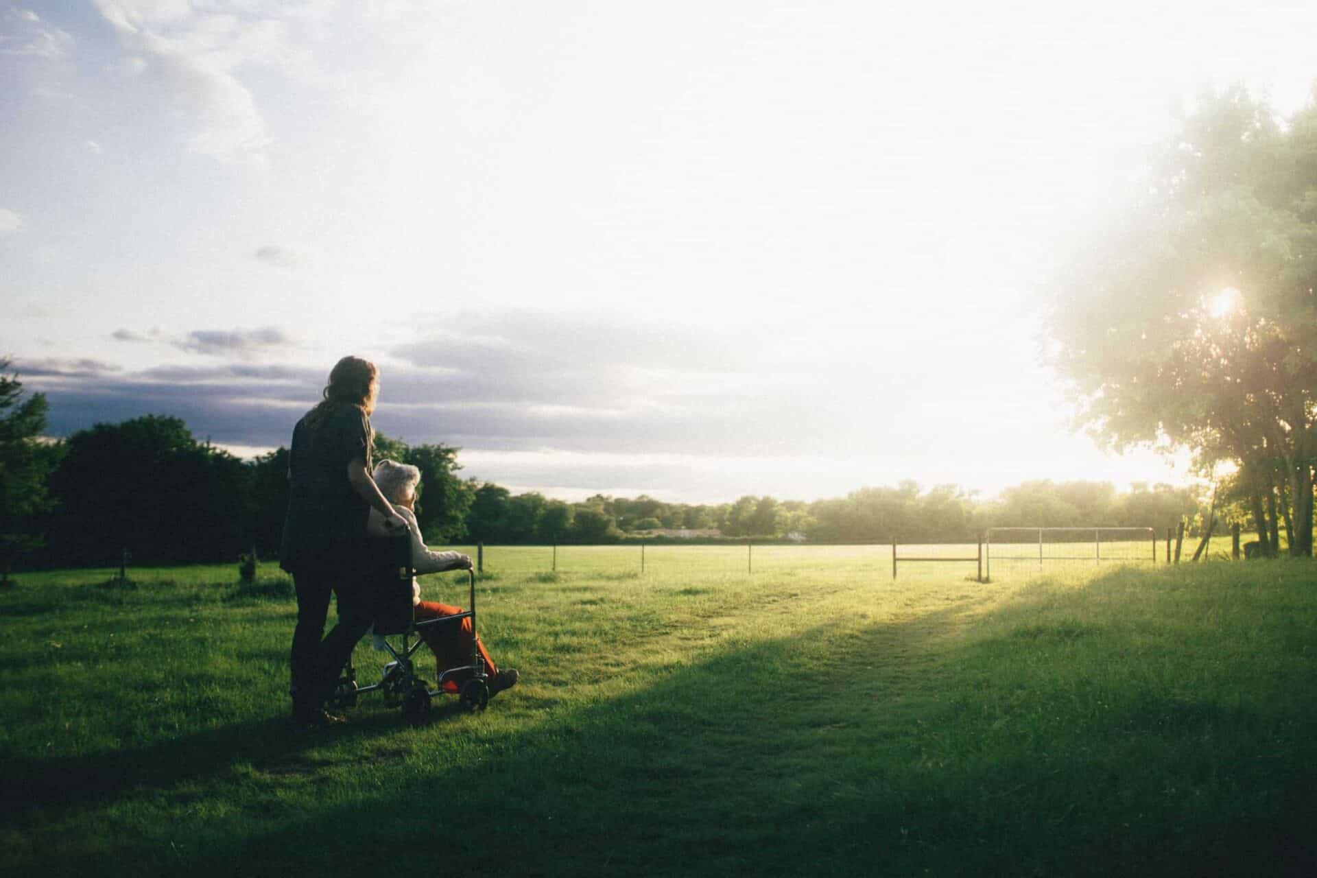 A serene evening walk happens: a person pushing a stroller along a grassy path near a fence with the warm glow of the setting sun filtering through the trees.