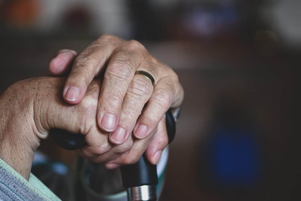 Close-up of an elderly person's hands clasped over the handle of a walking cane in a nursing home.