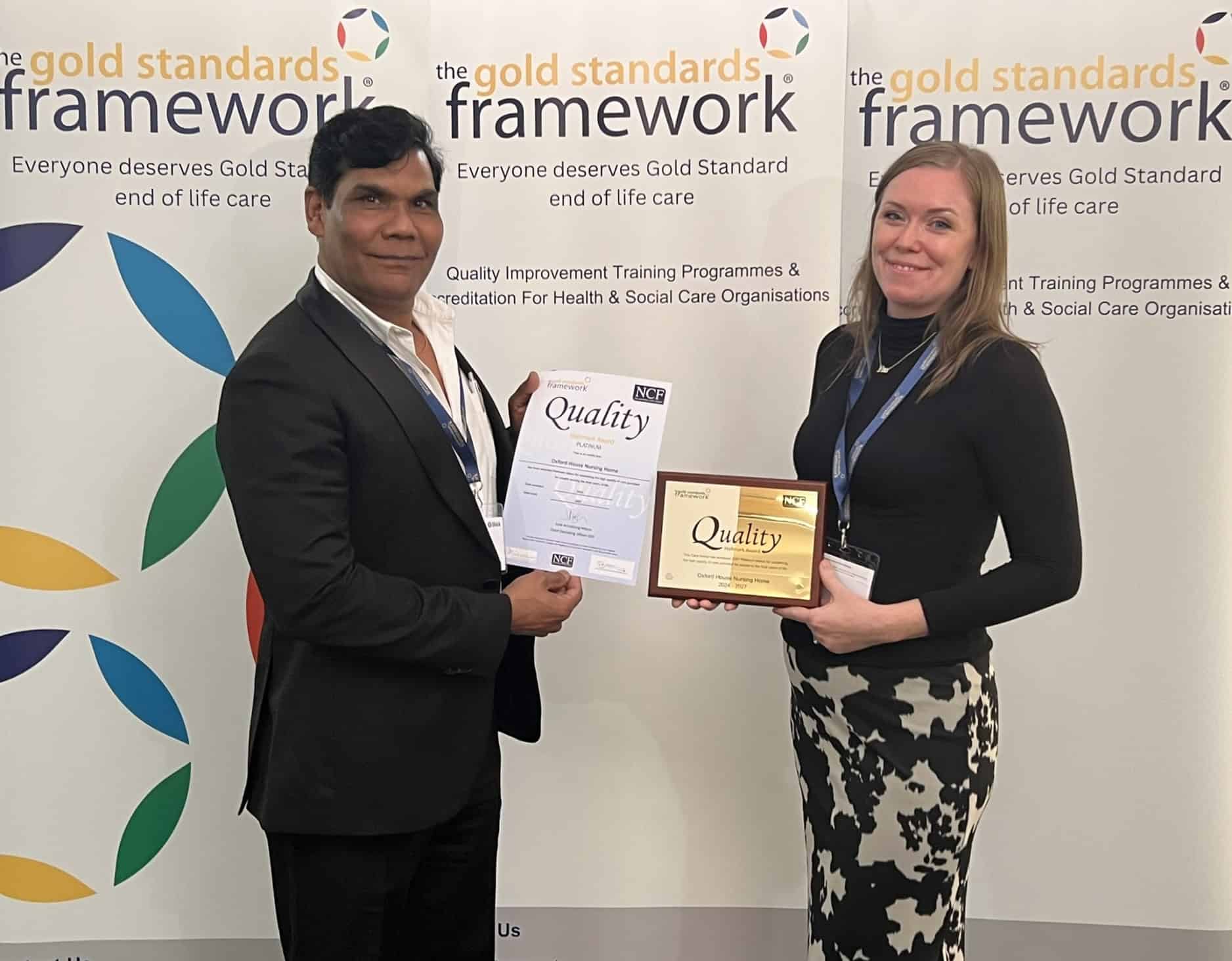 Two people stand smiling and holding an award and a certificate in front of a banner that reads, "The Gold Standards Framework," celebrating their commitment to gold standard end-of-life care.