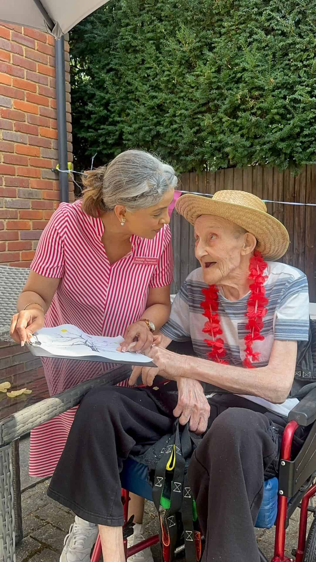 A woman in a pink striped dress shows a piece of paper to an elderly person in a straw hat and red lei, who is sitting in a wheelchair. They are outdoors by a brick wall and wooden fence, smiling at each other.