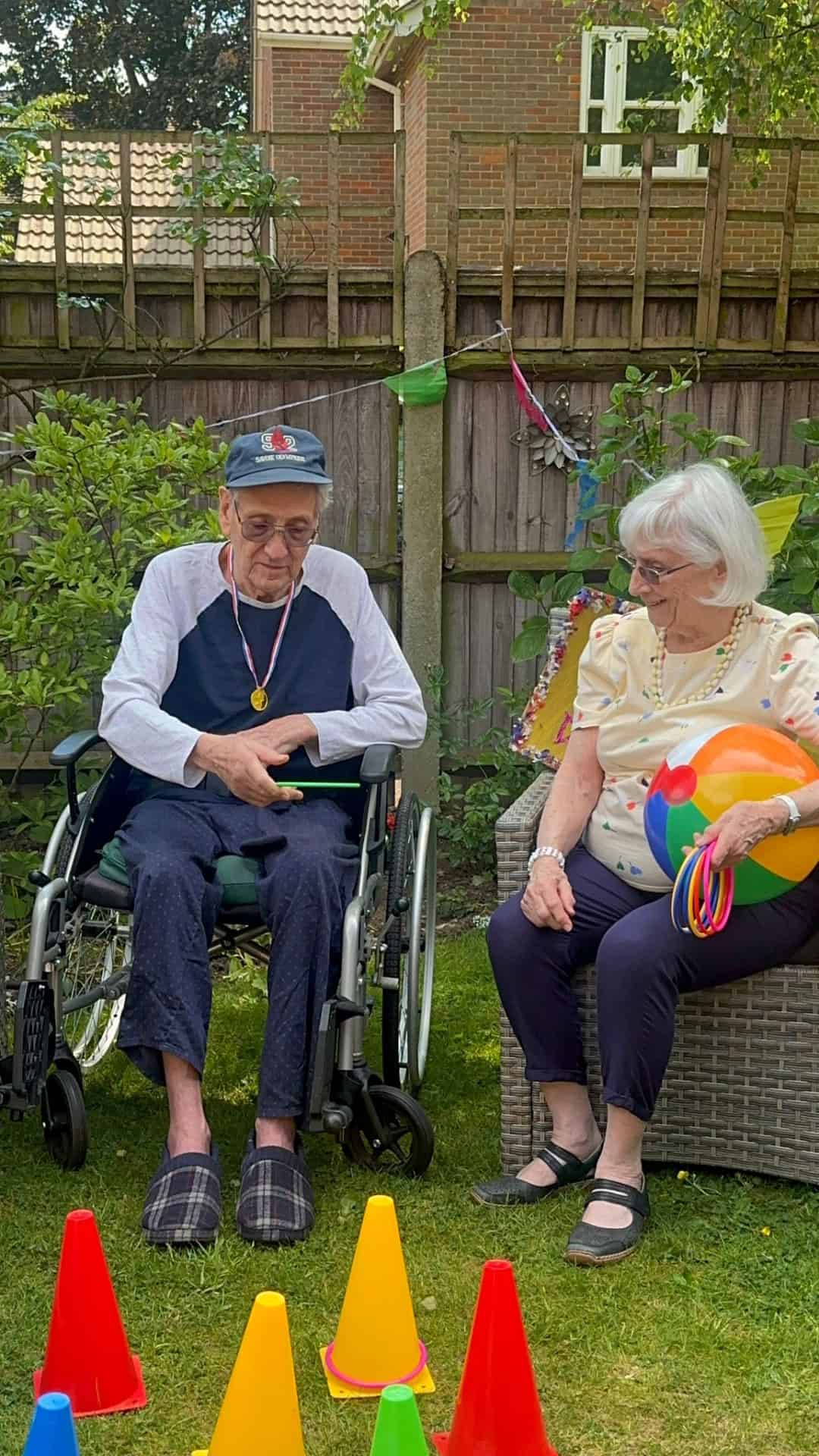 An elderly man in a wheelchair and an elderly woman sit outside in a garden. The woman holds a colorful ball and plastic rings. There are colorful cones arranged on the grass in front of them. Both appear to be enjoying a game.