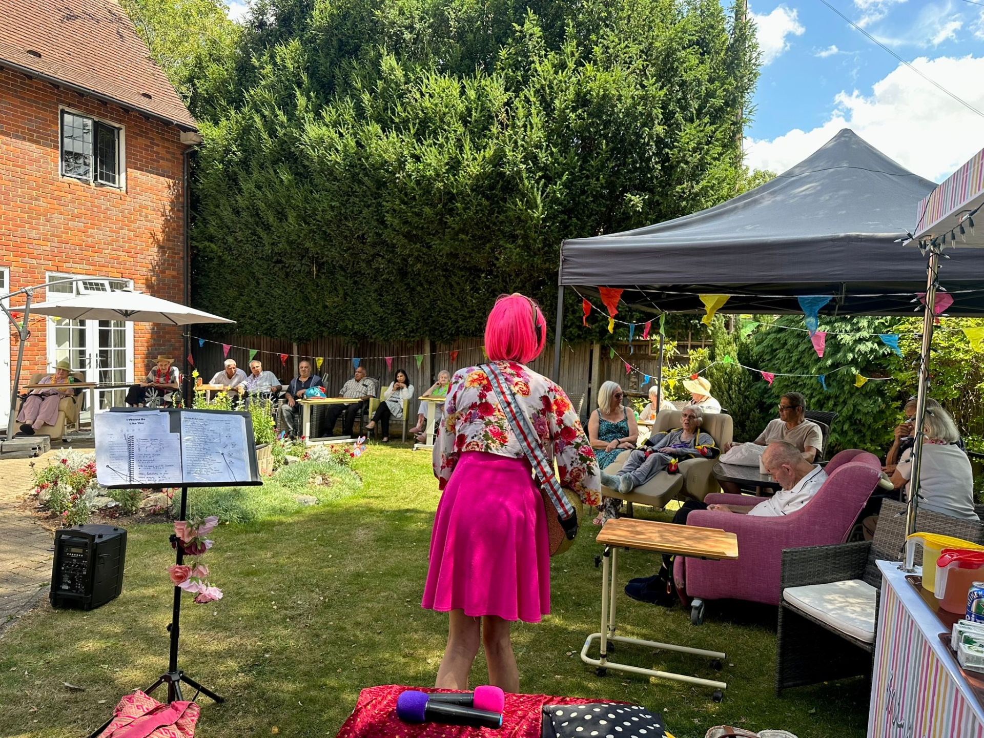 A woman with bright pink hair and a pink skirt stands in a garden, leading events for an audience seated under a canopy and on the lawn, surrounded by colorful bunting and greenery on a sunny day.