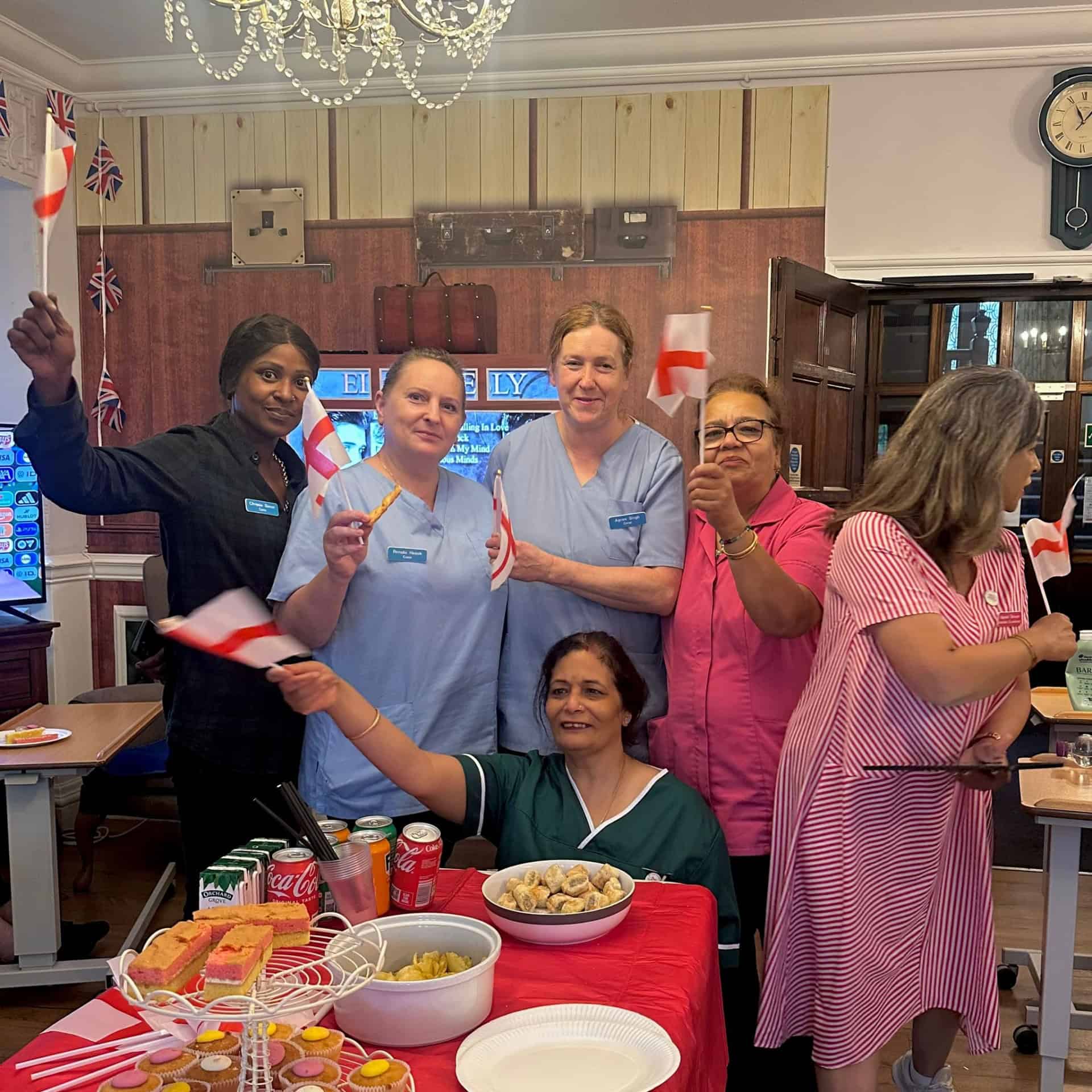 A dedicated team of five women, some in uniforms and others in casual clothes, smile and wave small English flags around a table with food and drinks in a decorated room. Another woman sits nearby, also waving a flag.