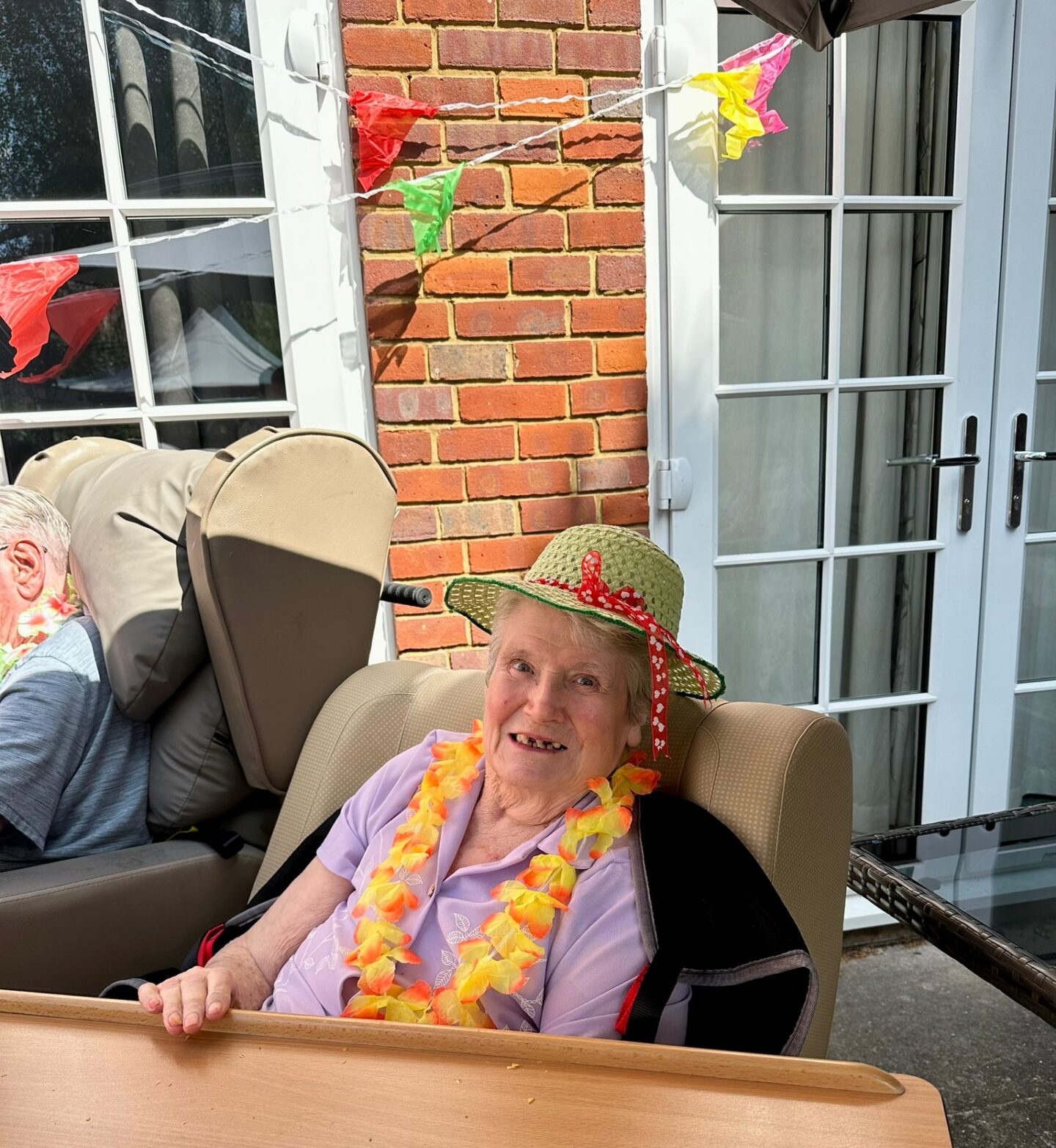 An elderly woman wearing a green hat and a colorful flower lei smiles while sitting at a table outdoors at Oxford House Nursing Home. Festive decorations, bunting, large windows, and a brick wall create a cheerful atmosphere behind her.